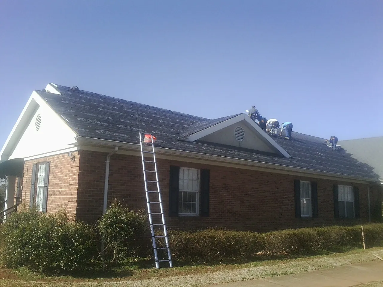 Crew tearing shingles off a brick ranch roof with a ladder set — Emergency Roof Repair work by our crew in Gilberton