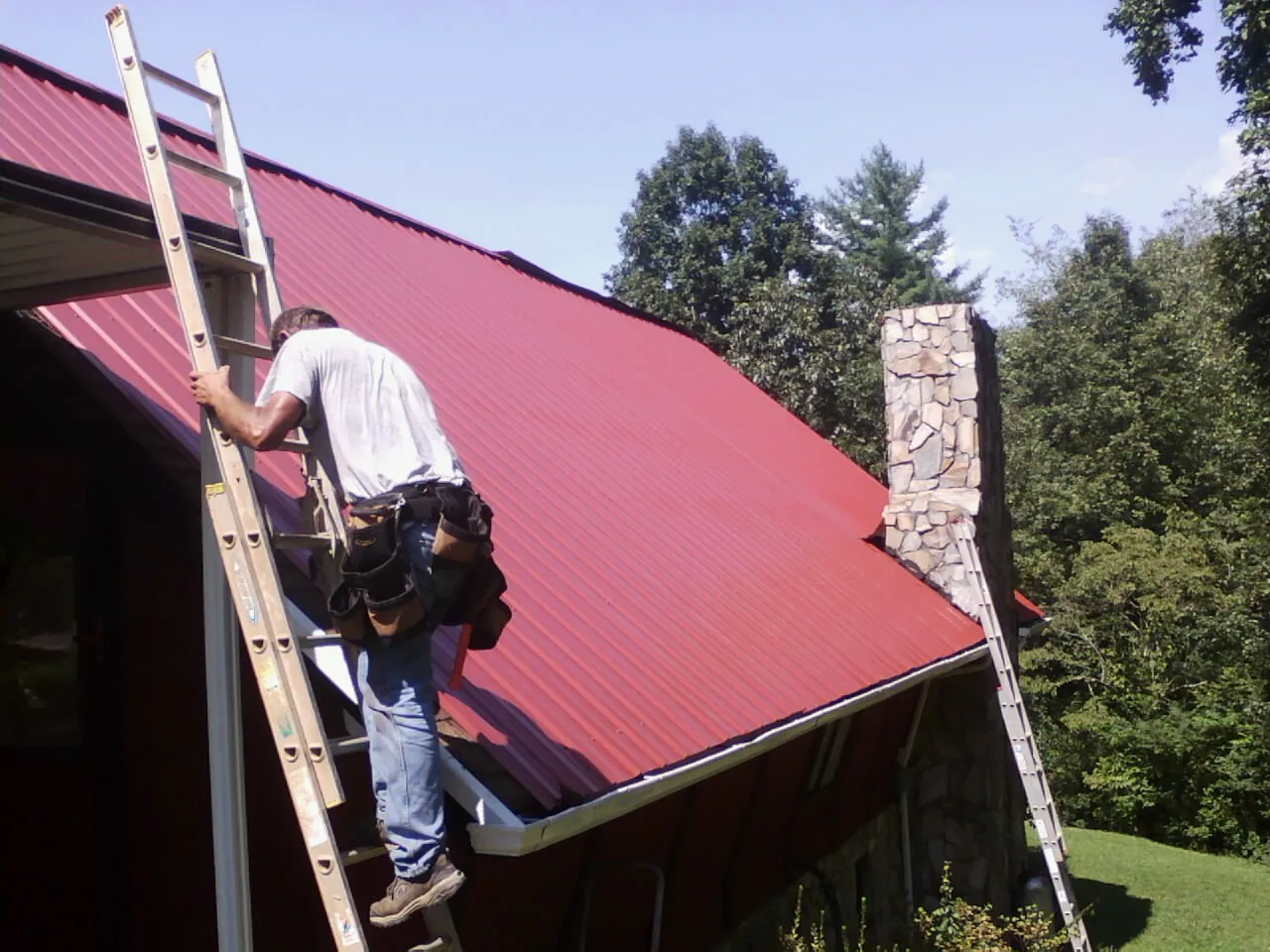 Roofer on a ladder beside a red metal roof and stone chimney — Soffit & Fascia Repair by Roofing Brothers in Gilberton
